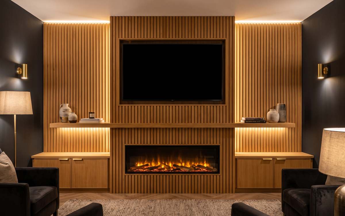 A fitted media wall in oak slatted panelling with a recessed television and a lit fire below it